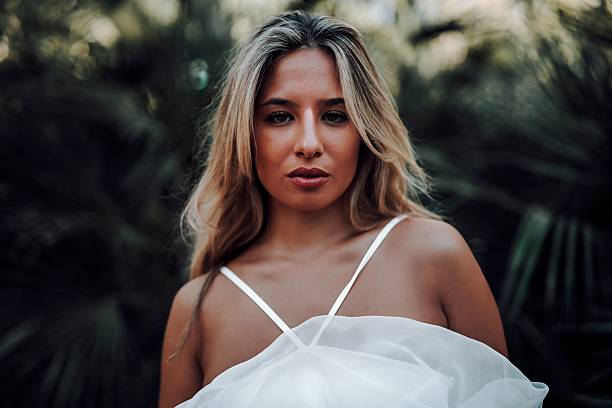 Portrait of a young woman with long blonde hair in a white dress, set against a blurred natural background.