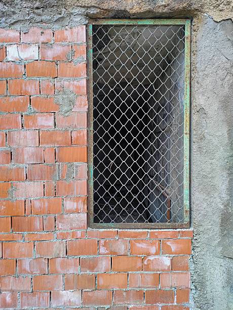 A brown brick wall with relief sides is held together with cement mortar. A window with an iron frame with a metal mesh rests against a concrete wall and ceiling.