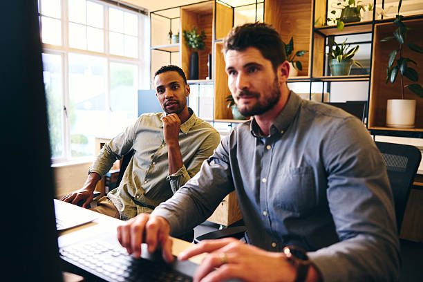 istock Two businessmen in office, one using desktop PC, the other watching with hand on chin, concentrating 2209001712