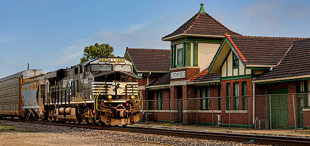 Railroad crossing signals with red and white striped arms flank a road leading to a grand brick courthouse in Richmond, Texas. The historic building's distinctive architecture stands against a bright blue sky with puffy white clouds. Lush green trees frame the scene, blending old and new elements in a quintessential small-town American landscape.