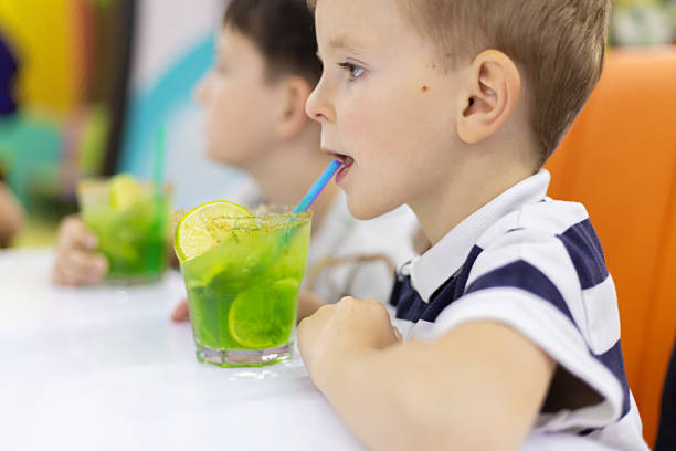 Child in striped shirt drinking bright green mocktail with lime slices and mint through a straw while sitting at a white table in an indoor setting