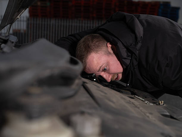 A concerned man stands in front of his broken-down car on the side of a car park. The car's hood is open, revealing the engine, while the man appears to be waiting for assistance. The setting is an overcast day, emphasising the feeling of isolation. This high-quality stock image captures themes of travel issues, car trouble, roadside emergencies, and the need for roadside assistance