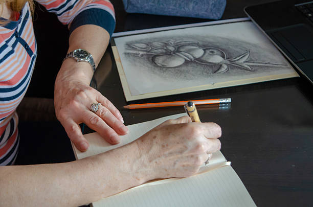 A dedicated senior woman sits at her table, carefully taking notes in her notebook as she follows an online course on her laptop, enjoying her free time away from work. With her blonde hair and casual attire, she is fully immersed in her creative process. Her focused expression shows how much she cherishes this peaceful moment of personal growth and artistic expression.