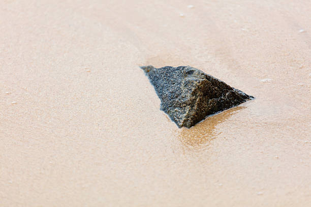 A rock is sitting on the sand. The rock is small and has a triangular shape. The sand is light brown in color