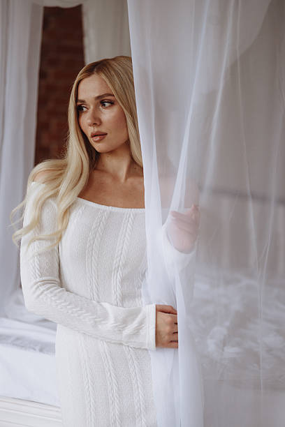 Front view of a young blonde woman in an off-shoulder white dress, stands on a white bed under a sheer canopy, looking at camera. Concept lifestyle, beauty, bedroom decor.