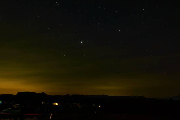A vast night sky with countless twinkling stars on a pitch black canvas. The horizon is quietly tinged with a mysterious light, perhaps the lights of a city or a sign of dawn. On the ground, small camping lights are scattered against the backdrop of the majestic mountain silhouette, telling the quiet lives of people living in the great outdoors. A tranquil and romantic night scene where the grandeur of the universe and the warmth of the earth intersect.