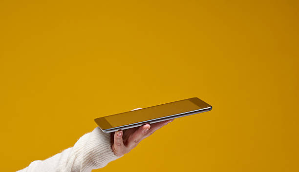 istock Hand wearing a white sweater holds a silver tablet against a solid yellow background. The tablet screen is blank and facing upwards, copy space 2209060765