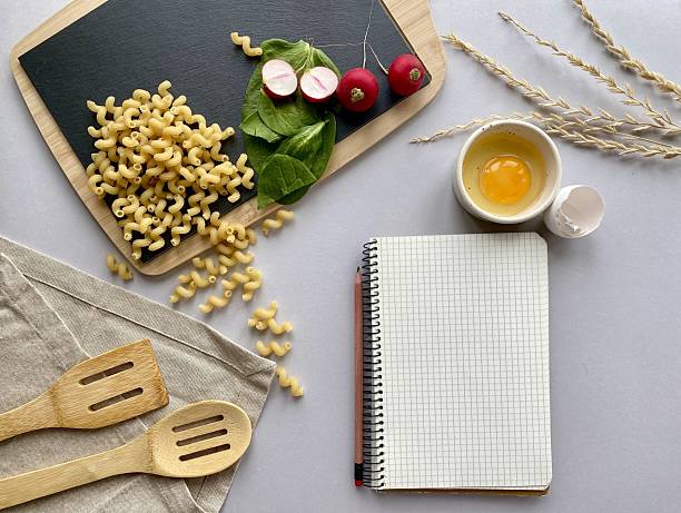 Pasta, radish, greens on the black board. Egg in the bowl, wheat and notebook on the grey background. Empty space for captions. Empty space for recipe. Cooking theme.