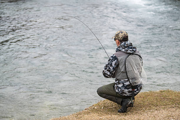 An angler wearing gray camouflage gear is fly fishing in a calm river surrounded by nature. The photo captures a tranquil moment of outdoor recreation, with the fisherman mid-cast in a serene natural setting.