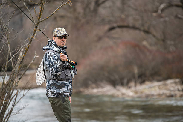 An angler wearing gray camouflage gear is fly fishing in a calm river surrounded by nature. The photo captures a tranquil moment of outdoor recreation, with the fisherman mid-cast in a serene natural setting.