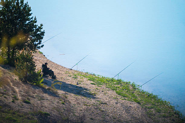 A lone fisherman is seen casting his fishing rod on a peaceful lake, surrounded by still water and natural scenery. The image captures the tranquility of recreational fishing in a serene outdoor setting.