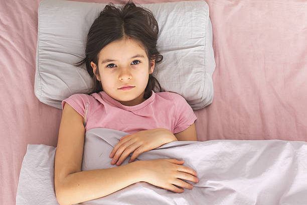 A sick girl lying in bed under a light blanket with a thermometer under her arm, looking tired and unwell. She rests on a pillow in a pink-themed bedroom, indicating fever and recovery, top view