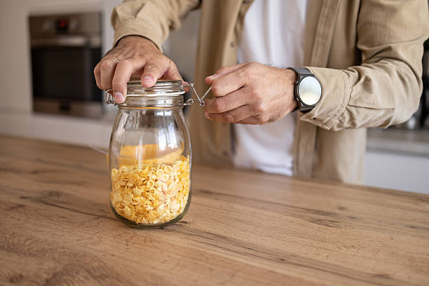 Man wearing a smartwatch closes a swing top jar filled with cornflakes on a wooden kitchen counter, preserving freshness for a healthy breakfast or snack option