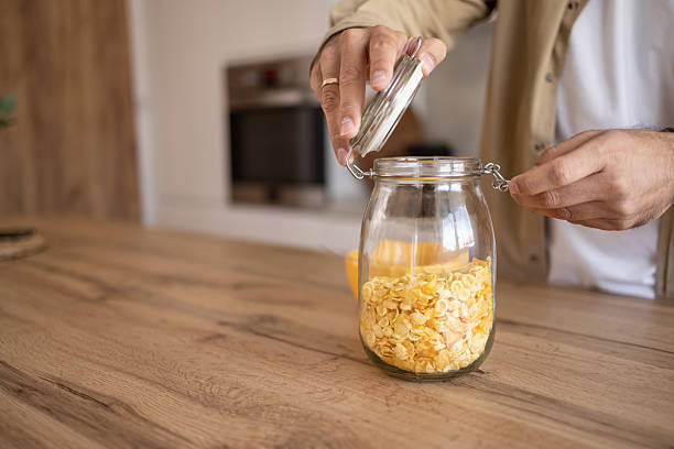 Man closing a glass jar filled with crunchy cornflakes on a wooden kitchen counter, creating a tidy and organized space for healthy breakfast options in a cozy home