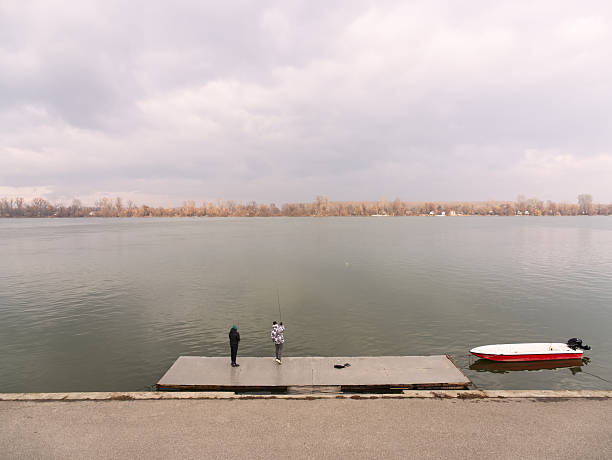 Belgrade, Serbia - January 12, 2025: Tranquil Danube river scene with two people on a concrete dock, red boat nearby, overcast sky. Zemun, Belgrade