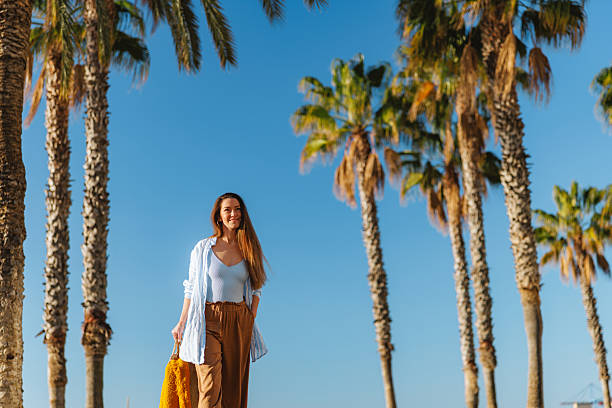 A young woman enjoys a carefree walk under palm trees at a beach in Malaga, Spain. The sunny day and clear blue sky enhance the feeling of relaxation and vacation joy.