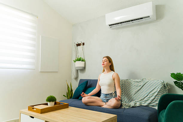 istock Peaceful woman meditating on sofa with air conditioning unit refreshing room 2209077571