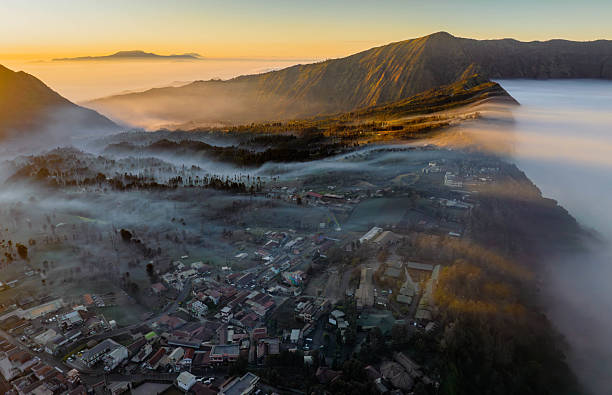 Witness the enchanting beauty of Bromo town in Indonesia as the morning volcanic mist gently drifts over the houses and landscape. This stunning aerial view captures the serene and almost otherworldly atmosphere of the village nestled near the majestic Mount Bromo. Experience the magical interplay of light, fog, and the silhouette of the surrounding mountains in this breathtaking scene