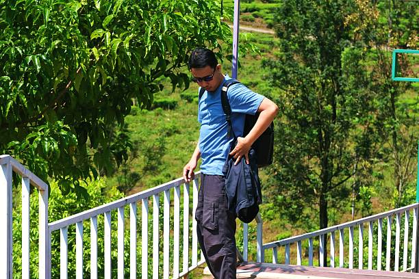 A young adult in a white polo shirt and jeans stands casually against a lush green ivy wall, hands in pockets. They exude confidence and ease, with a direct gaze and relaxed posture.