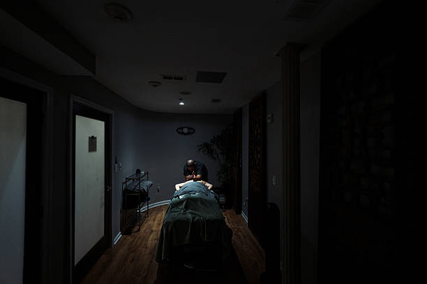 Wide shot of an unrecognizable woman laying face down on a massage table in a dimly lit private treatment room at a luxury wellness center and spa as a  Black female massage therapist massages her back, with spotlight lighting illuminating the client's back.