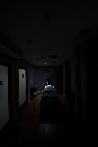 Wide shot of an unrecognizable woman laying face down on a massage table in a dimly lit private treatment room at a luxury wellness center and spa as a  Black female massage therapist massages her back, with spotlight lighting illuminating the client's back.