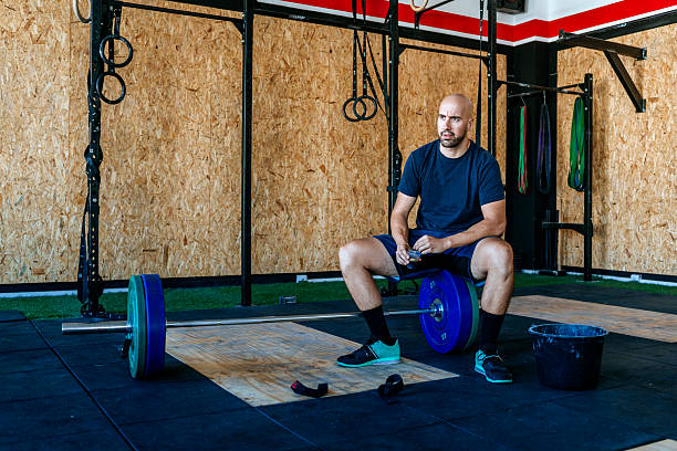 Man drinking coffee sitting on a dumbbell in the gym.