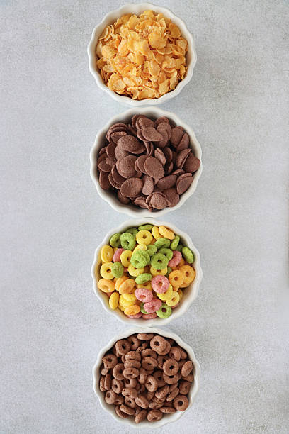 Image of row of four bowls of dry breakfast cereal, corn flakes, chocolate flavoured crunchy wheat shells, chocolate flavoured wheat hoops, multicoloured, fruit flavoured, crunchy, O-shaped cereal, mottled grey background, elevated view