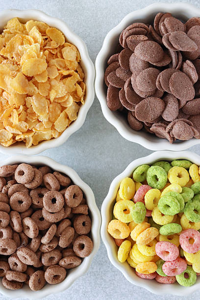 Close-up image of four bowls of dry breakfast cereal, corn flakes, chocolate flavoured crunchy wheat shells, chocolate flavoured wheat hoops, multicoloured, fruit flavoured, crunchy, O-shaped cereal, mottled grey background, elevated view