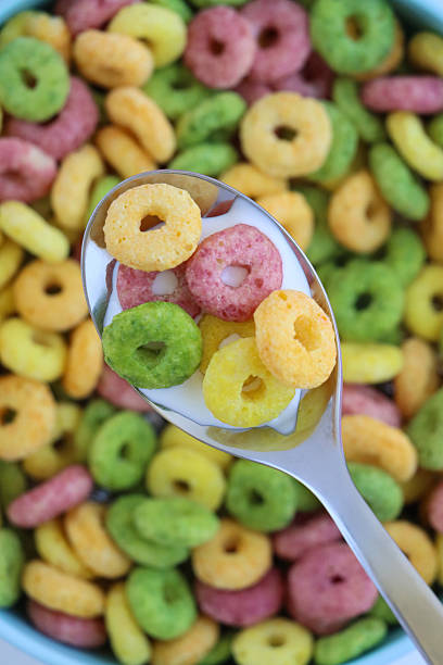 Full frame image of stainless steel spoonful of yellow, red, green and orange fruit flavoured crunchy O-shaped cereal, blue bowl of breakfast cereal with milk, made with wheat, oat, maize flours, sugar, elevated view