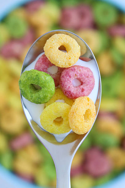 Full frame image of stainless steel spoonful of yellow, red, green and orange fruit flavoured crunchy O-shaped cereal, blue bowl of breakfast cereal with milk, made with wheat, oat, maize flours, sugar, elevated view