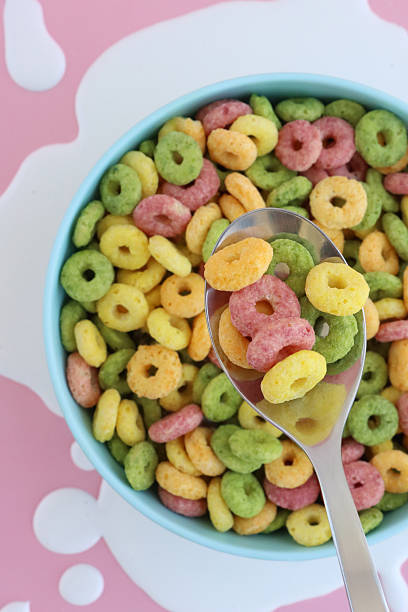 Full frame image of blue bowl of dry breakfast cereal with metal spoon surrounded by spilled milk, yellow, red, green and orange, fruit flavoured, crunchy, O-shaped cereal, made with wheat, oat, maize flours, sugar, pink background, elevated view