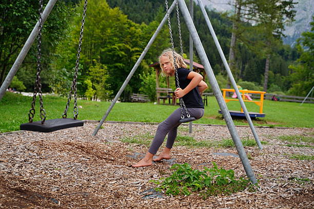 Child sitting on a swing set in a green park, looking pensive. Barefoot, relaxed in outdoor setting. Surrounded by greenery, trees, and recreational structures on a summer day
