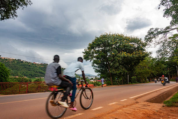 A moment of shared transportation in Kigali: two people ride a bicycle on a road with other vehicles and pedestrians nearby. The image conveys movement. The backdrop features a cityscape on a hill under a cloudy sky. This image is for editorial use.
