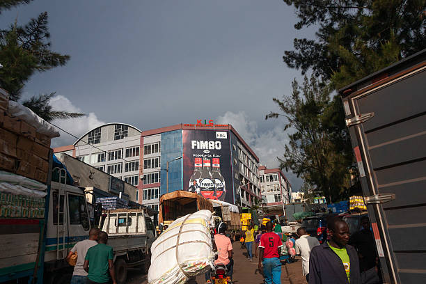 This editorial photograph captures a busy urban environment in Kigali City. Trucks laden with goods, motorcycles carrying passengers and cargo, and pedestrians fill the street. A prominent building with \