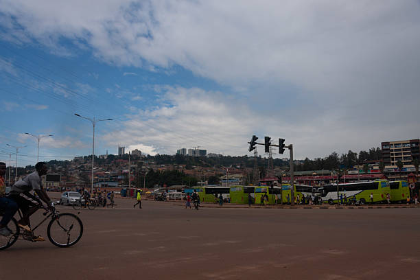 A wide shot captures a bustling urban intersection in Kigali, Kigali City, Rwanda, with cyclists navigating the road. Several large green and white buses are present, along with other vehicles and pedestrians. The background shows a densely built hillside under a sky with mixed clouds and patches of blue. Editorial use only.