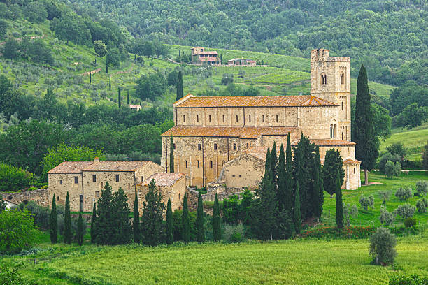 The magnificent Benedictine Abbey of Sant'Antimo stands serenely amid the verdant Tuscan landscape, its distinctive bell tower and honey-colored stone walls surrounded by elegant cypress trees and sprawling vineyards. This historic monastic complex near Montalcino presents a timeless scene against the backdrop of rolling hills, embodying the perfect harmony between spiritual heritage and the natural beauty of the Italian countryside.