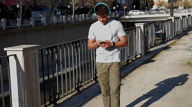 Young man enjoying a sunny day while walking along a river, listening to music through headphones and using his smartphone to stay connected and entertained