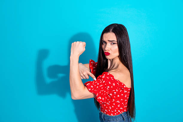 istock Confident young brunette woman pointing to her flexed bicep in a red top against a solid blue background 2209135035