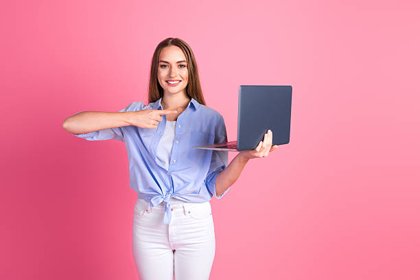 istock Young woman holding a laptop while pointing towards it on a vibrant pink background, emphasizing casual style 2209136700