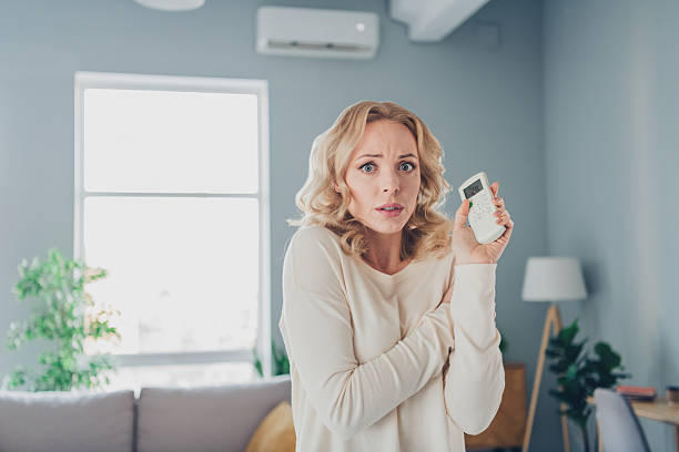istock Young blonde woman in a cozy living room holding a temperature remote controller while expressing a cold and uncomfortable feeling indoors 2209137632