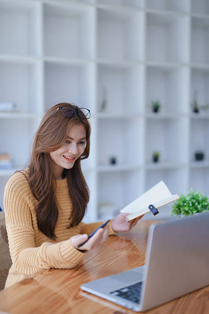 istock Smiling young woman engaged in a joyful online e-learning video call, gestu while holding a book with headphones on a wooden table nearby 2209139273
