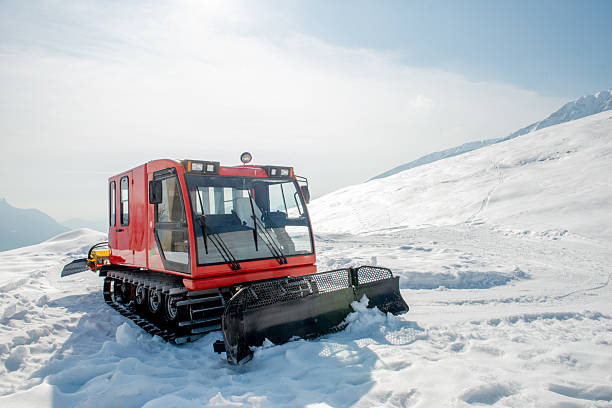 A snow grooming machine is parked on a snowy landscape, ready to maintain ski slopes. Surrounding mountains glisten in the sunlight on a clear day.