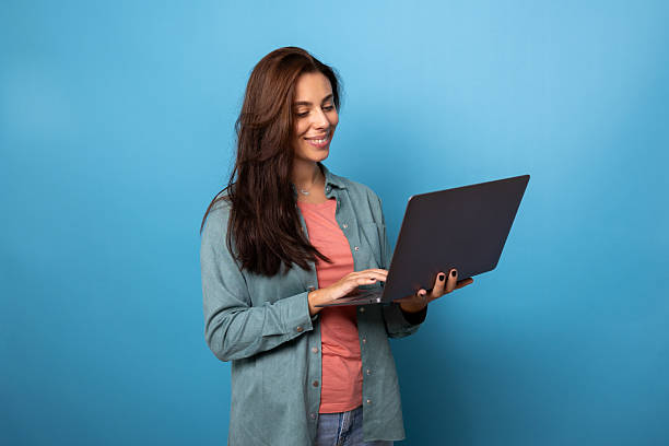 istock Young woman smiling and typing on a laptop against a blue backdrop 2209150867