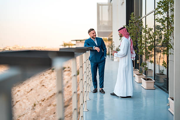 Professional portrait of young Middle Eastern businessman in traditional thobe and Western executive in navy suit engaging in conversation on modern high-rise office balcony overlooking desert landscape.