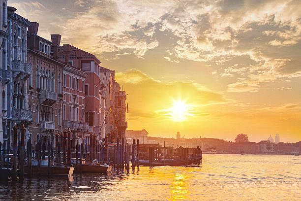 A scenic view of the lagoon in Venice at sunrise, with traditional gondolas docked along historic buildings. The warm sunlight reflects on the water, creating a golden glow that enhances the romantic atmosphere of the city.
