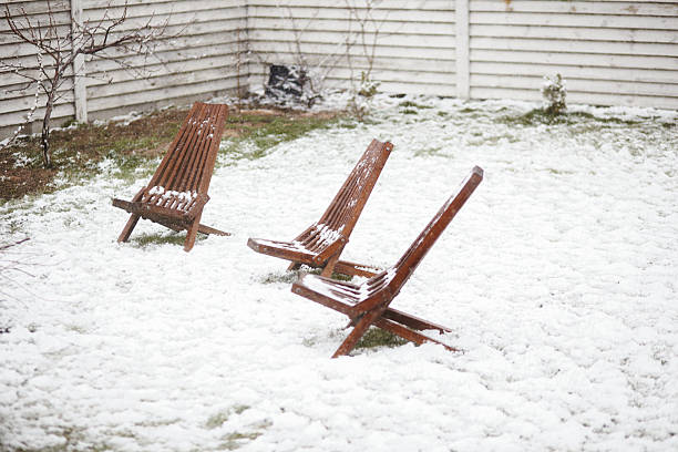 Three empty wooden chairs covered in snow in a corner of a backyard, symbolizing sudden weather change and stillness.