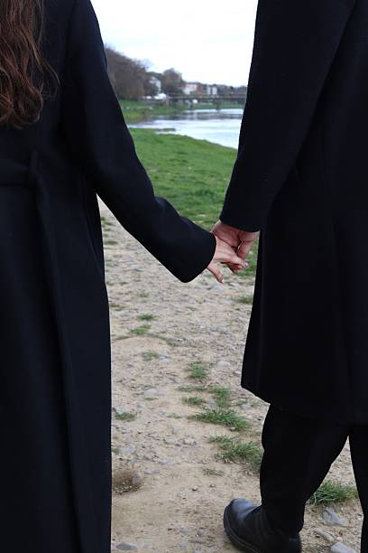 istock A romantic couple walks hand in hand along a riverside path, dressed in elegant black coats. Captured from behind, the image symbolizes love, unity, and peaceful moments in nature. 2209172525