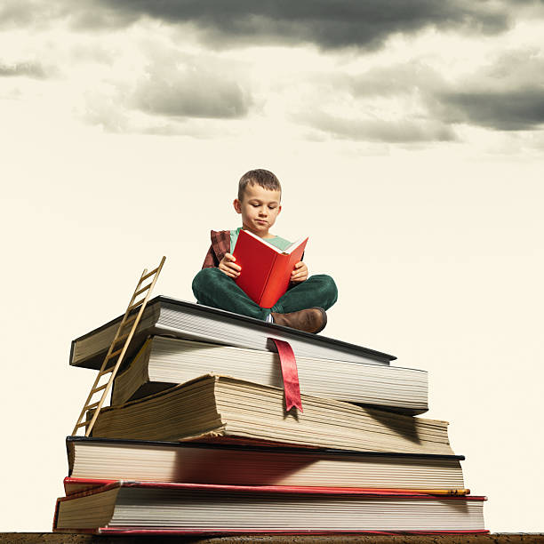 A boy holding a book sits atop a stack of books.