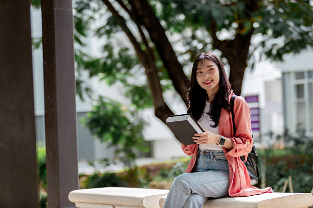istock Young Asian student sitting on a bench in a university campus, holding a book and smiling at the camera, enjoying outdoor study time 2209182247