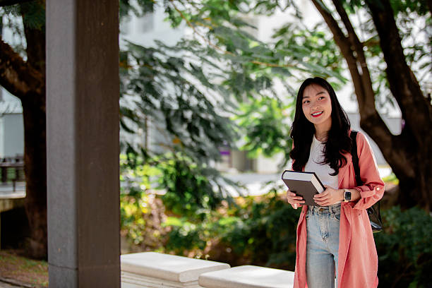istock Young Asian student smiling at the camera while holding a book on a university campus, surrounded by greenery and natural light 2209182254